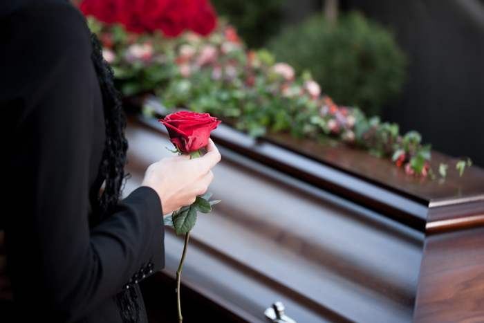 Bild: Rose wird in der Hand gehalten vor einem Sarg, welches im Hintergrund steht