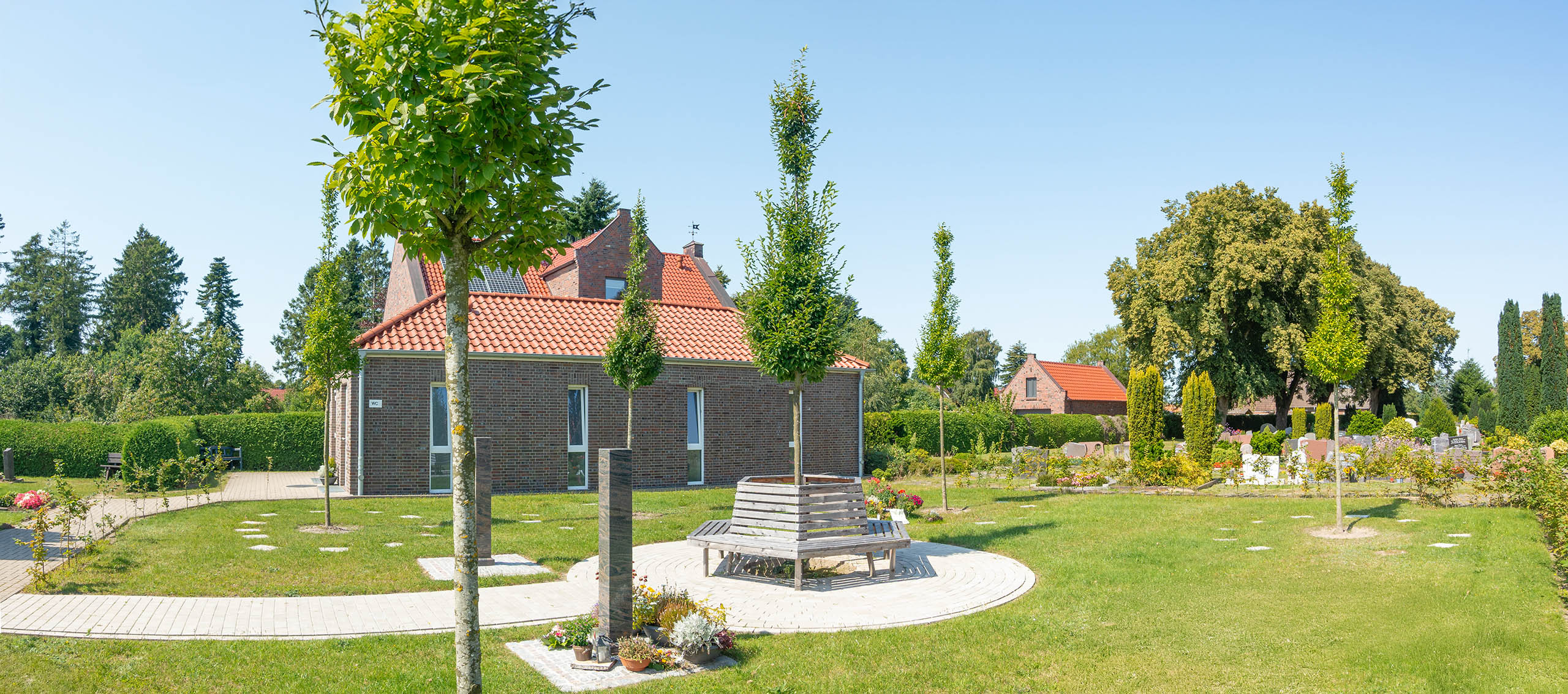 Überblick über den Baumhain auf dem Friedhof Schortens. Es sind Jungbäume zu sehen und eine Sitzgelegenheit mittig im Bild sowie 2 Stelen.